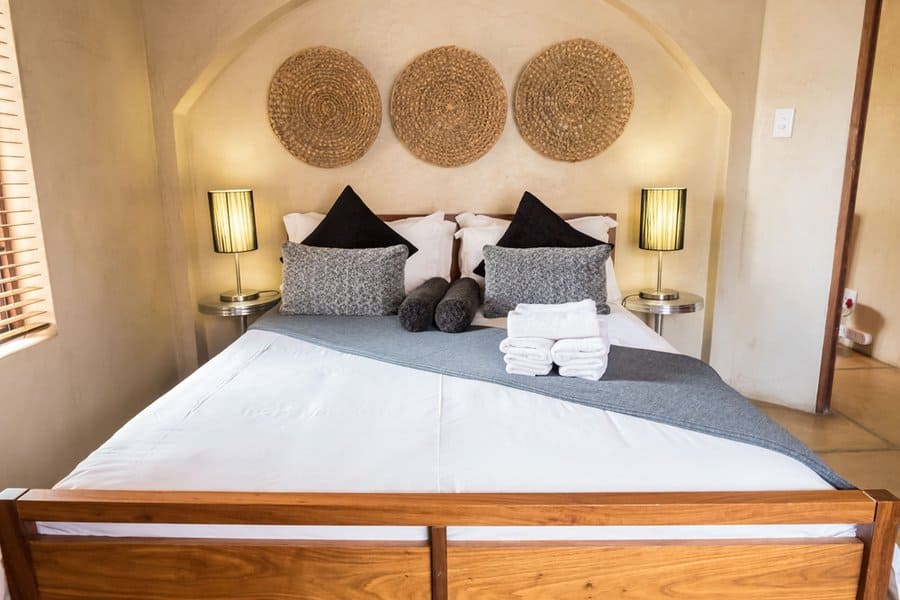 Cozy bedroom with a wooden bed, decorative pillows, lamps, and woven wall art above the headboard; towels are neatly placed on the bed