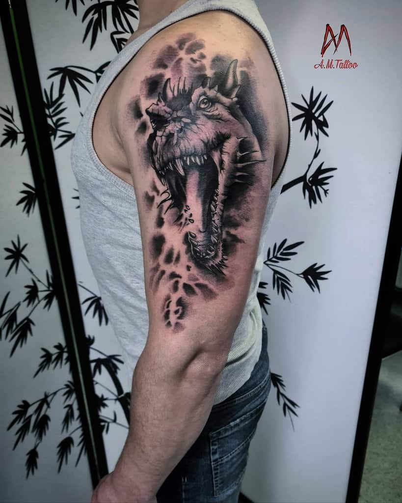 Intense upper arm tattoo featuring a fierce dragon's head with an open mouth, detailed in black and grey shading with a smoky background