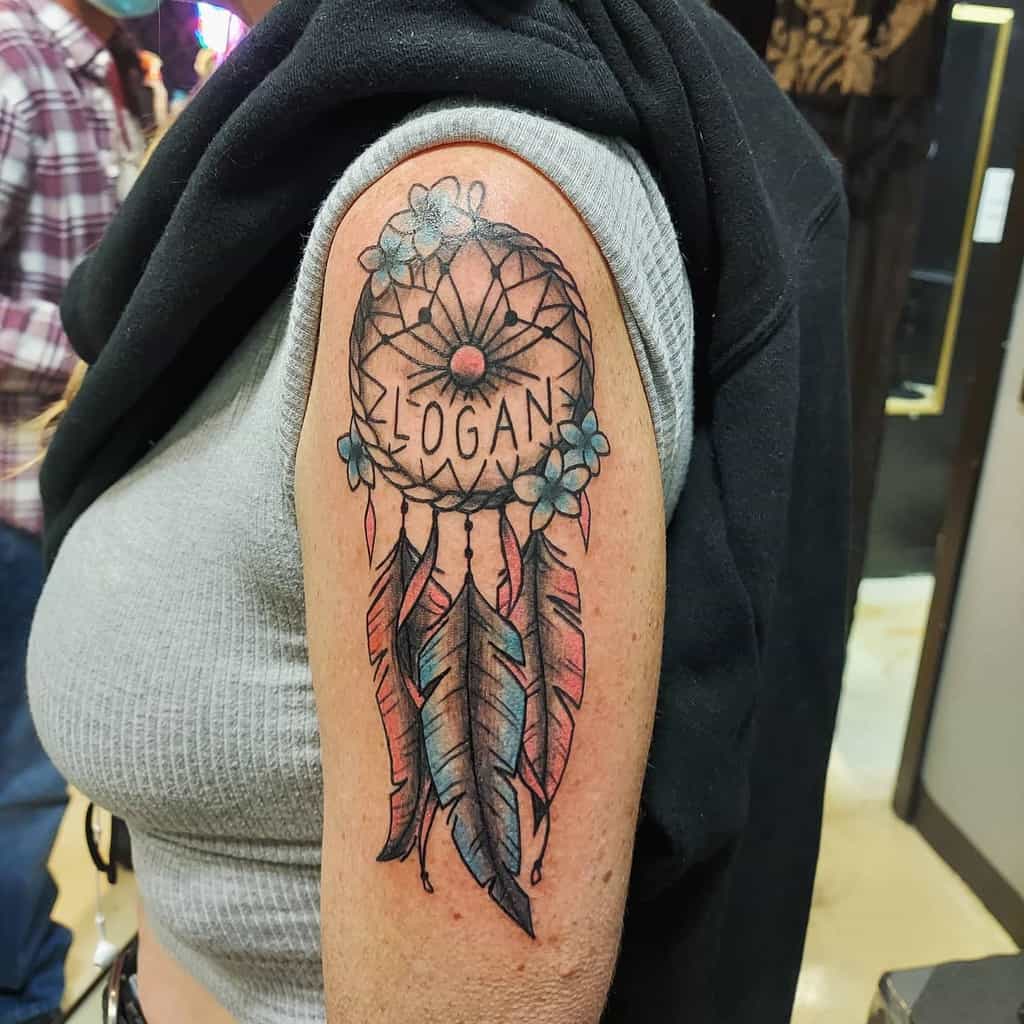 Dreamcatcher tattoo with the name "Logan," featuring blue flowers and feathers on upper arm
