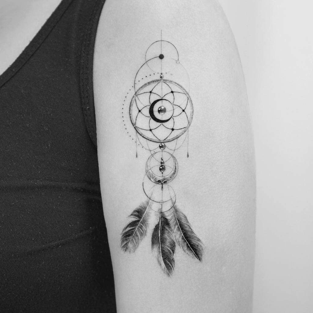 A black and white dreamcatcher tattoo with feathers on an upper arm