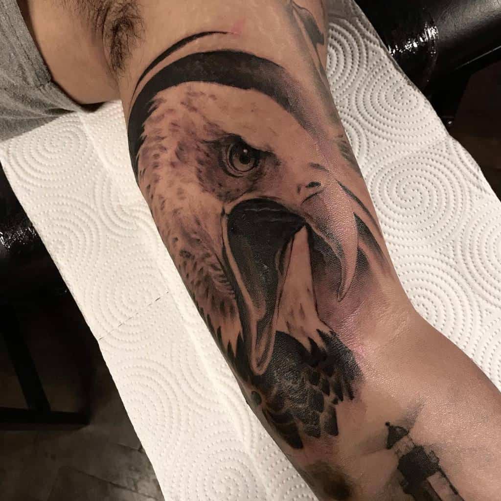 A detailed black and gray tattoo of an eagle's head on an arm, with a lighthouse at the bottom
