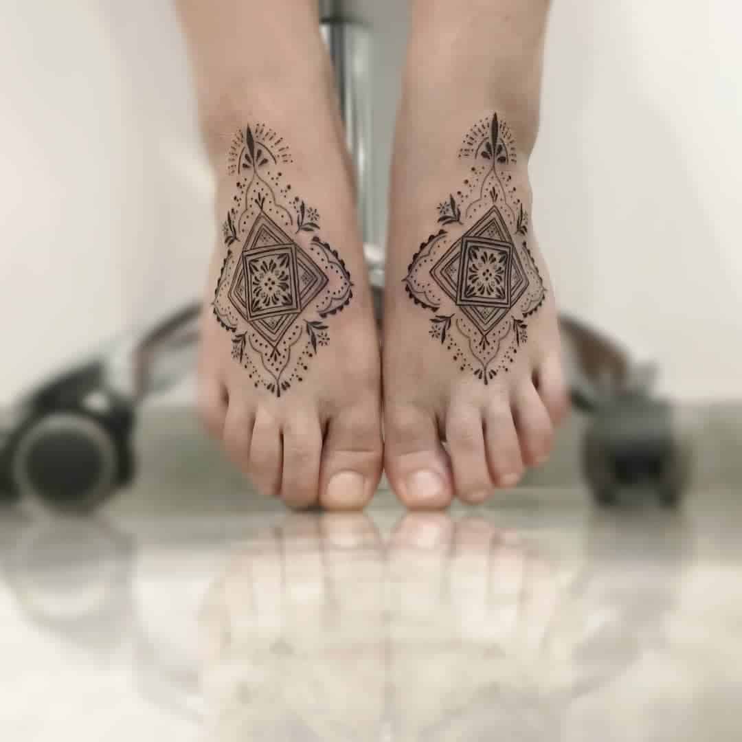 Two feet with intricate henna designs on top, resting on a reflective floor with a chair wheel visible in the background