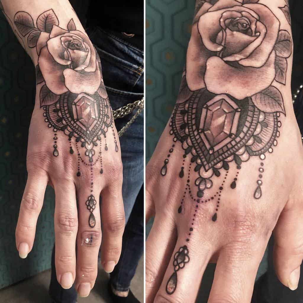 Hand tattoo featuring a detailed rose with leaves and intricate ornamental design with diamond gemstone and teardrop beads 
