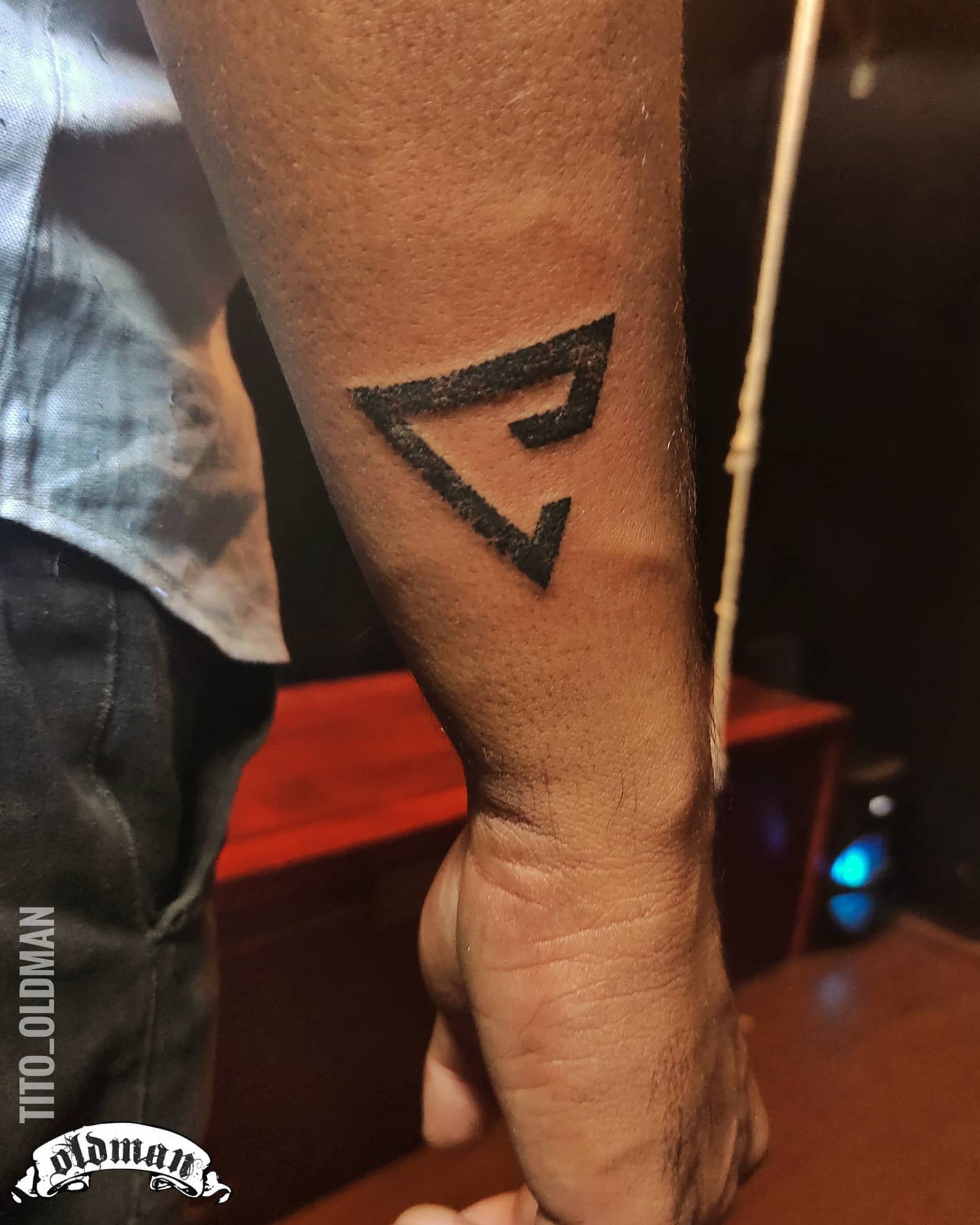 Forearm tattoo featuring a geometric triangle with a stylized 'E' inside in black ink