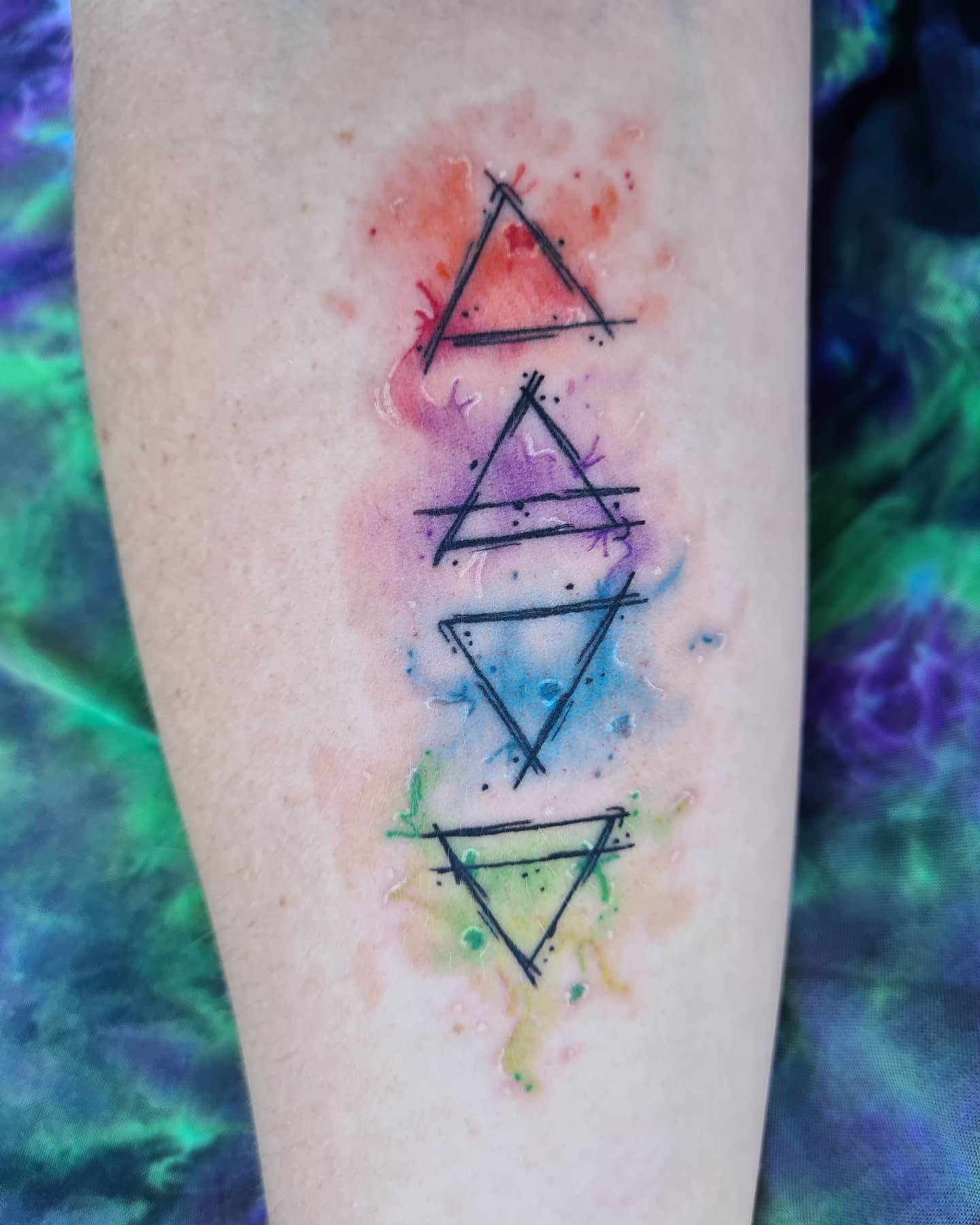 Forearm tattoo of four elemental symbols in black on a watercolor background