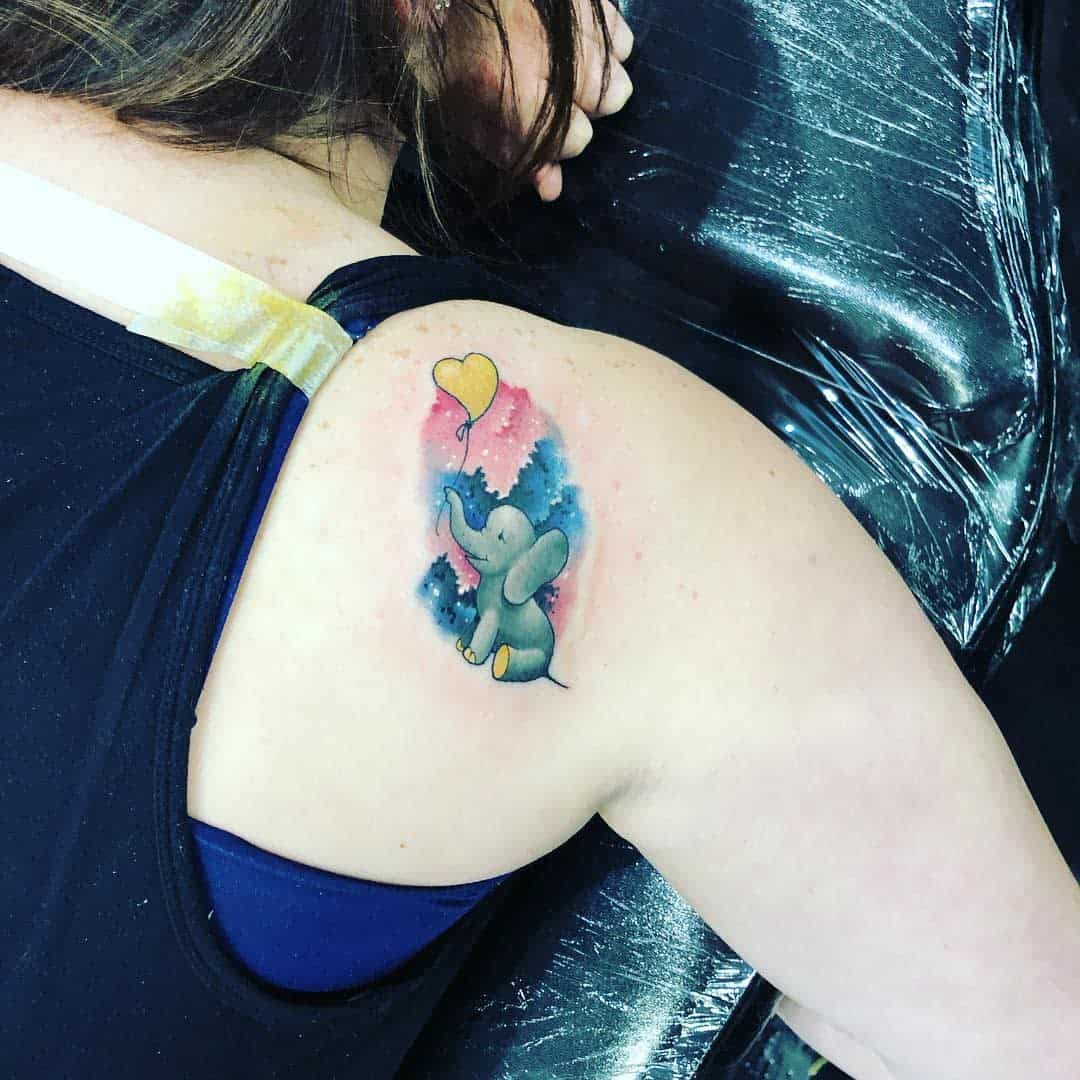 A tattoo of a small elephant holding a yellow heart balloon on a shoulder, with a pink and blue background