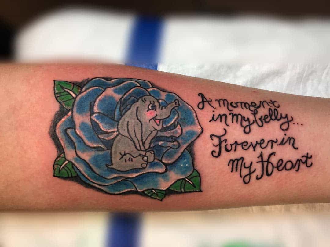 Tattoo of a baby elephant inside a blue rose with text: "A moment in my belly... Forever in my heart" on an arm