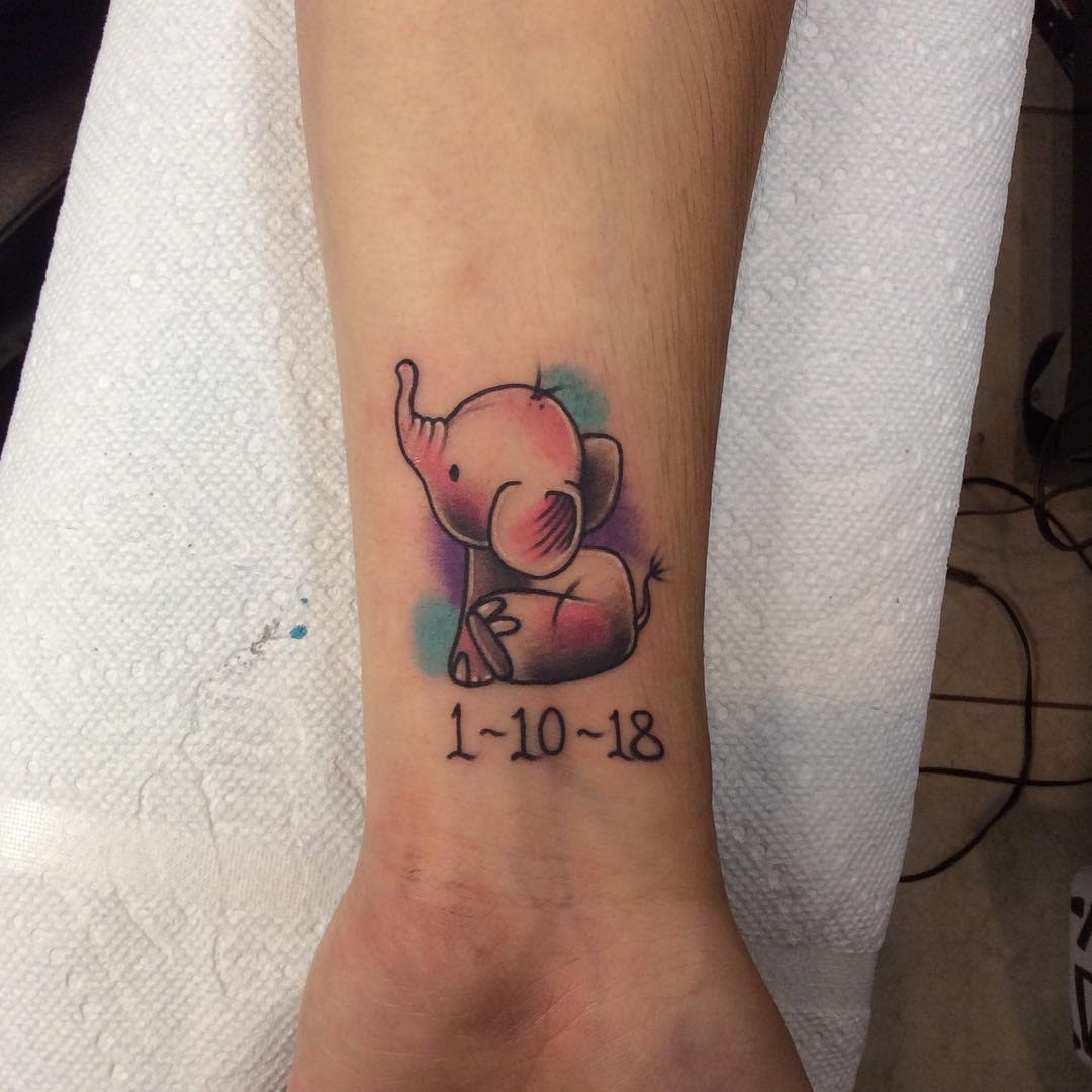 Tattoo of a cute baby elephant with date "1-10-18" on a forearm, colored with pastel shades