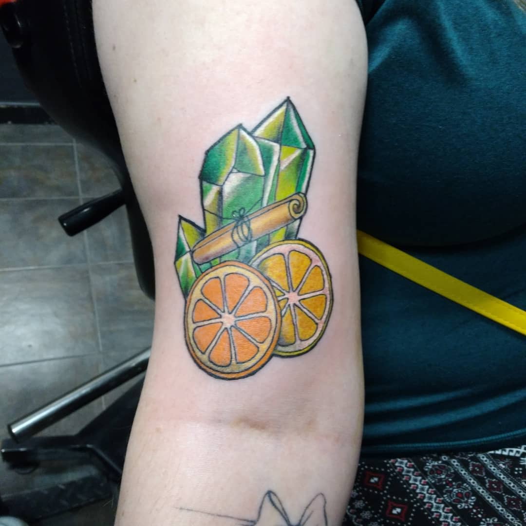 Tattoo of green crystals, a cinnamon stick, and orange slices on an upper arm