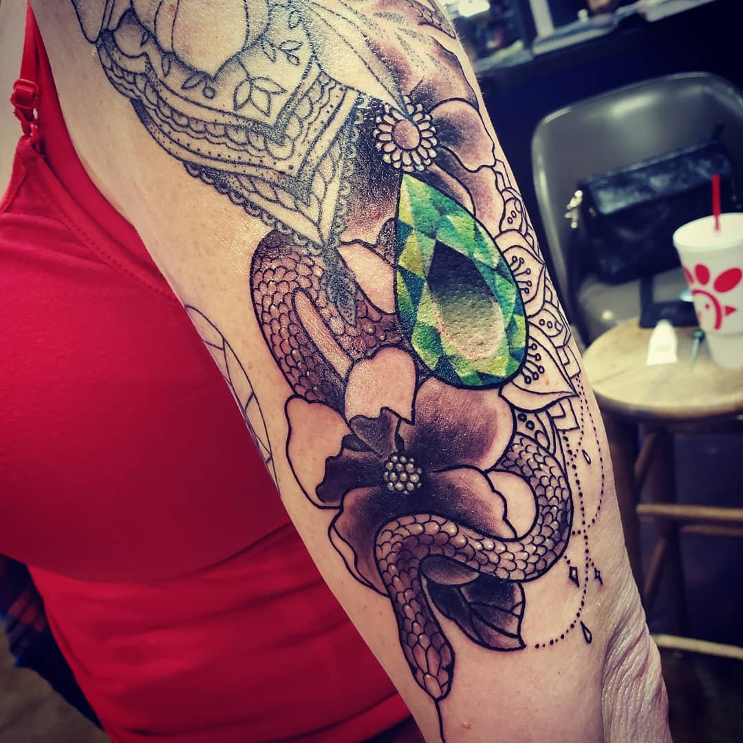 Arm tattoo featuring a snake, flowers, and a large green gemstone design