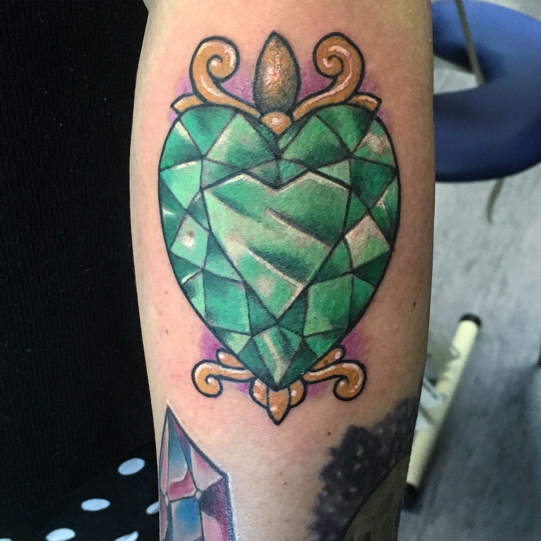 Tattoo of a green, gem-like heart with ornate gold details on an arm