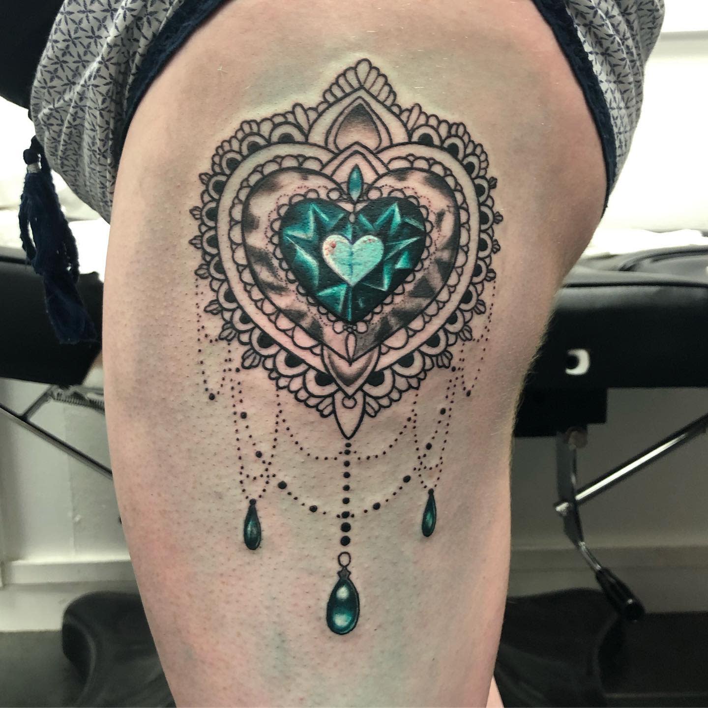 Intricate tattoo of a heart-shaped gem on a thigh surrounded by ornate patterns and dangling beads