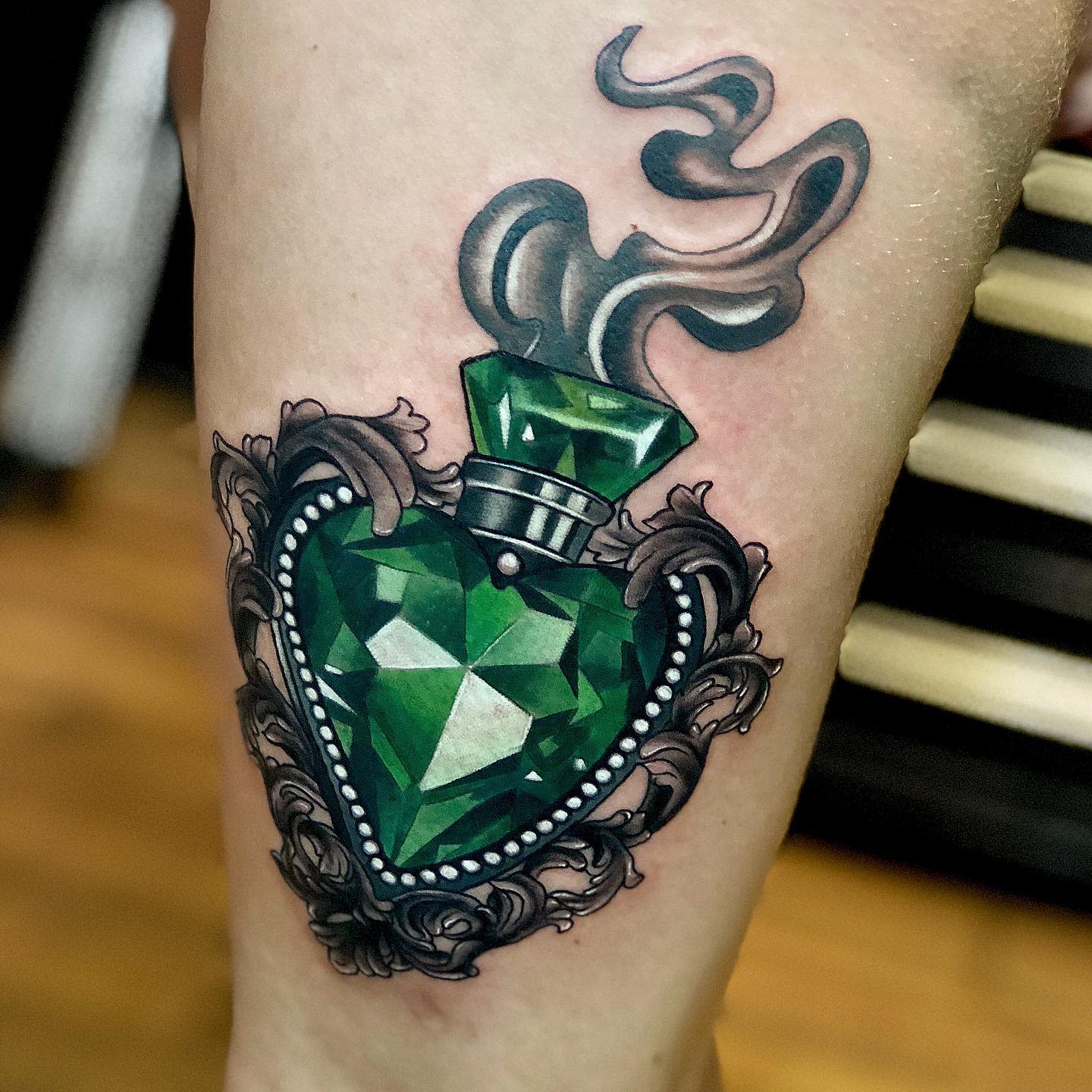 Tattoo of a heart-shaped emerald emitting smoke set in an ornate antique frame