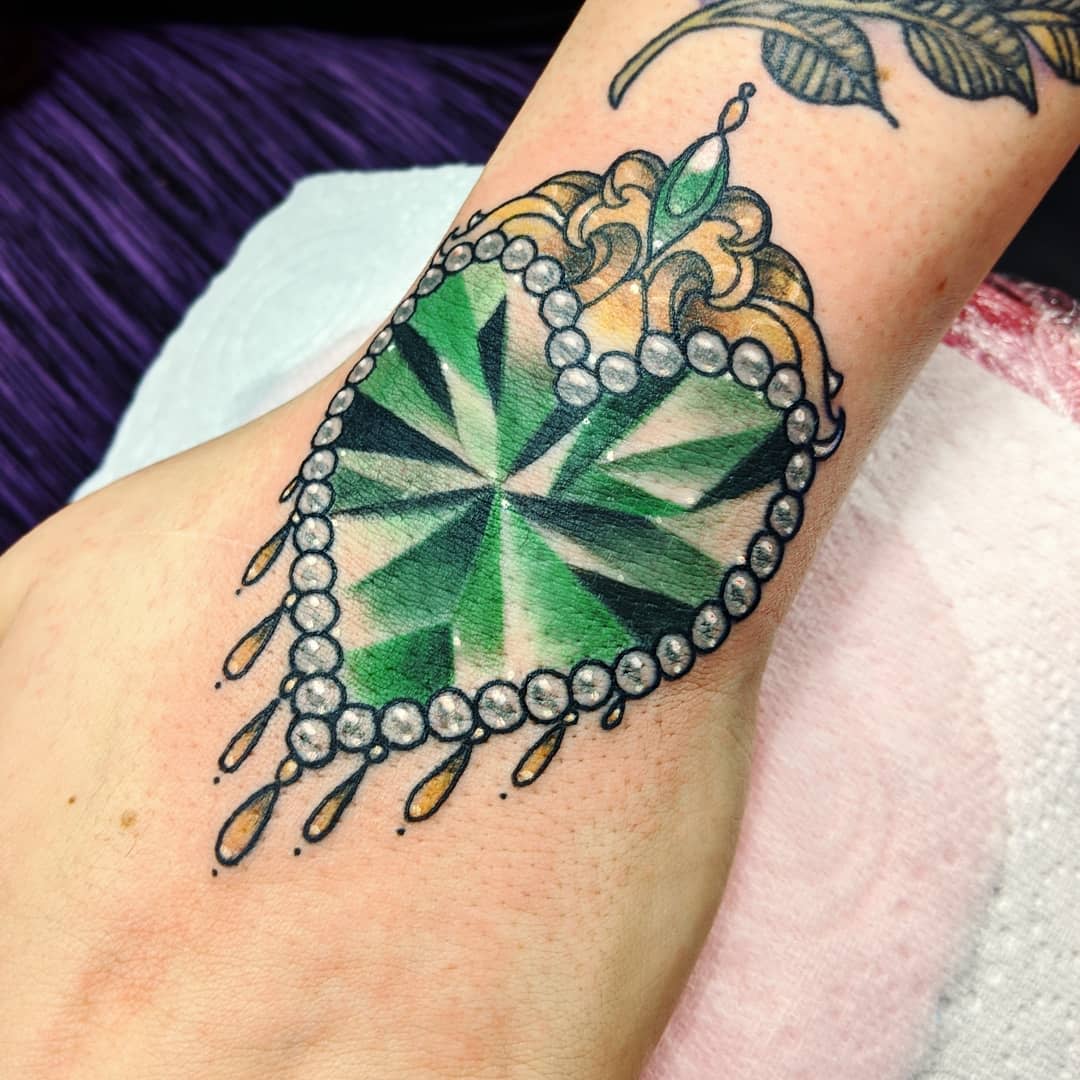 Heart-shaped emerald tattoo with beaded border and decorative top on a wrist