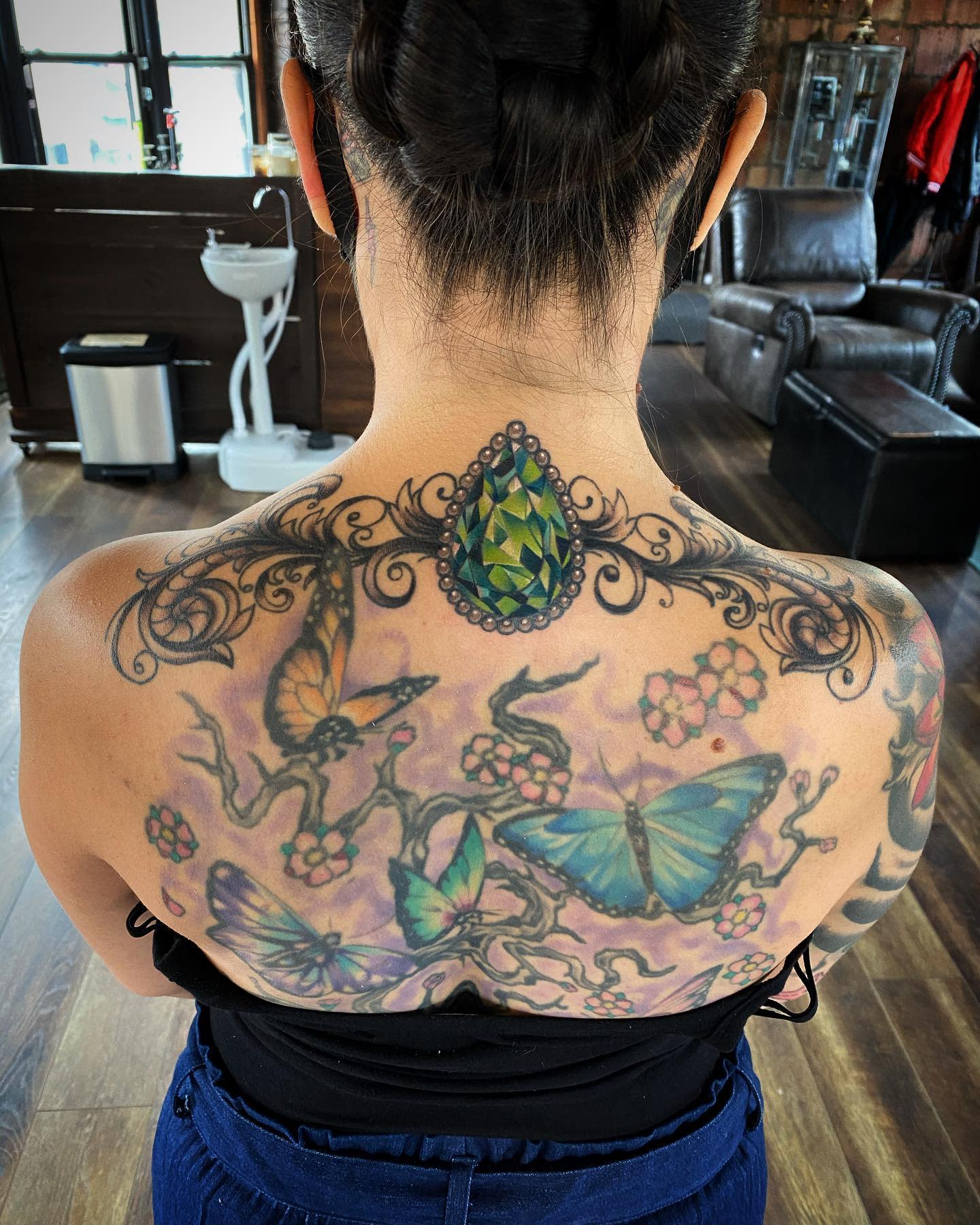 Woman showcasing a colorful back tattoo with butterflies, flowers, and a green gemstone