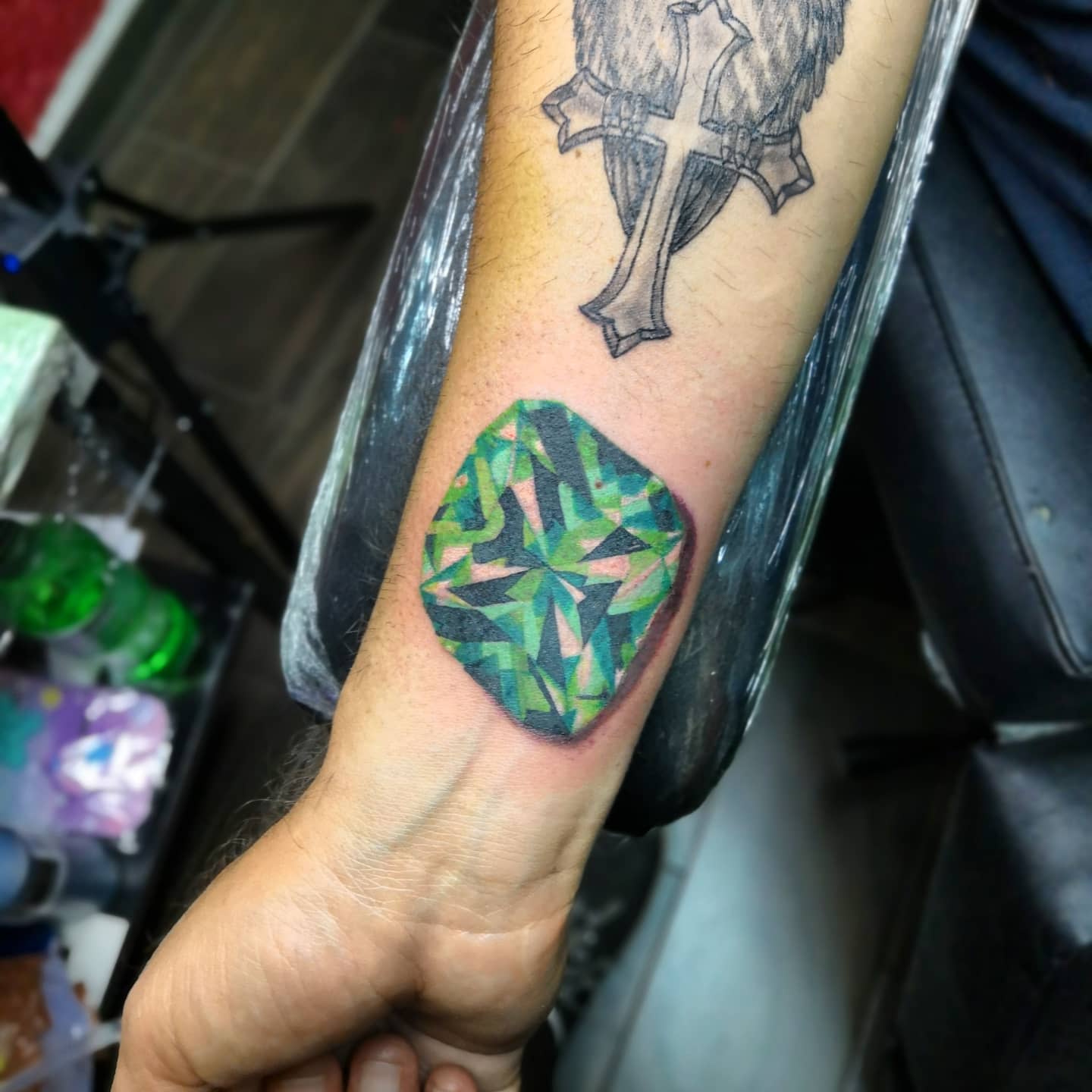 Forearm tattoo featuring a green geometric gemstone below a black and grey cross symbol