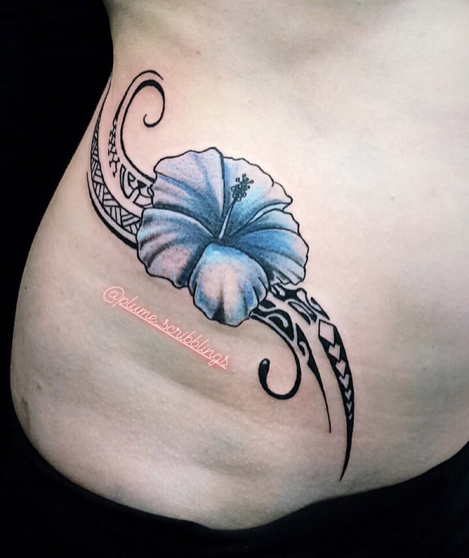 A blue hibiscus tattoo on the side of a torso adorned with intricate black Polynesian tribal designs