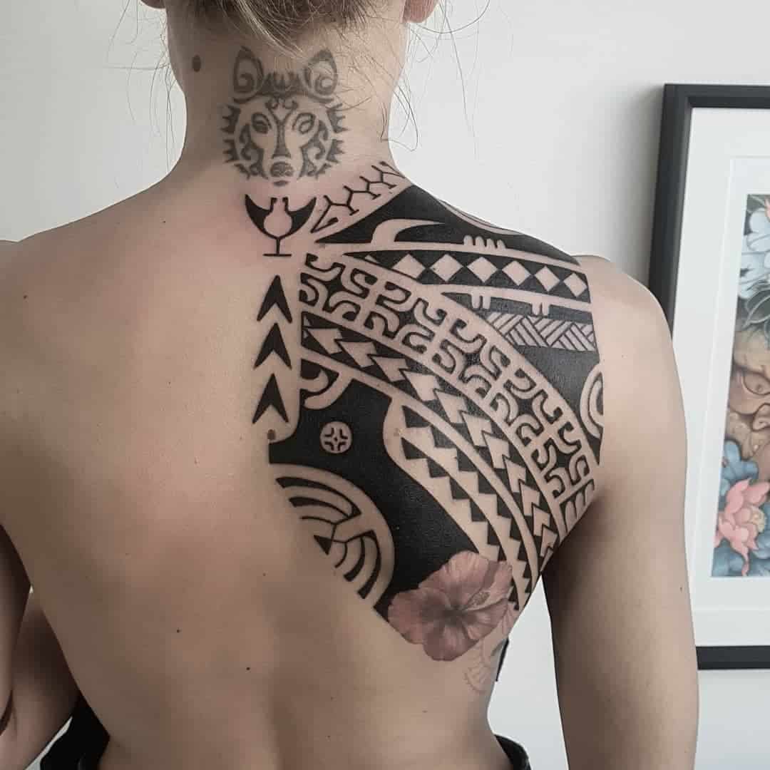 Individual with intricate Polynesian tribal-style tattoos covering their back and a wolf tattoo on their neck, viewed from behind
