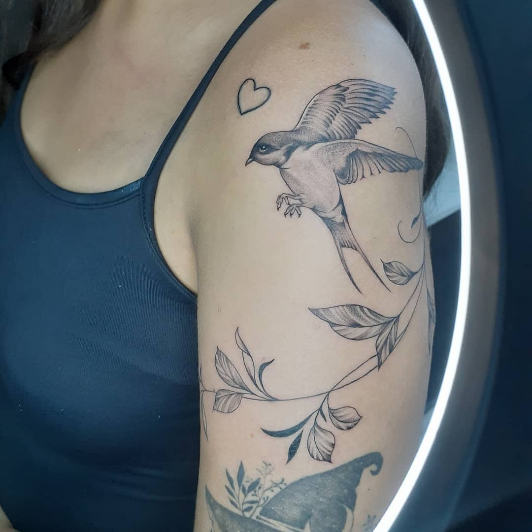A bird tattoo in flight on a woman's upper arm, featuring leaves and a small heart
