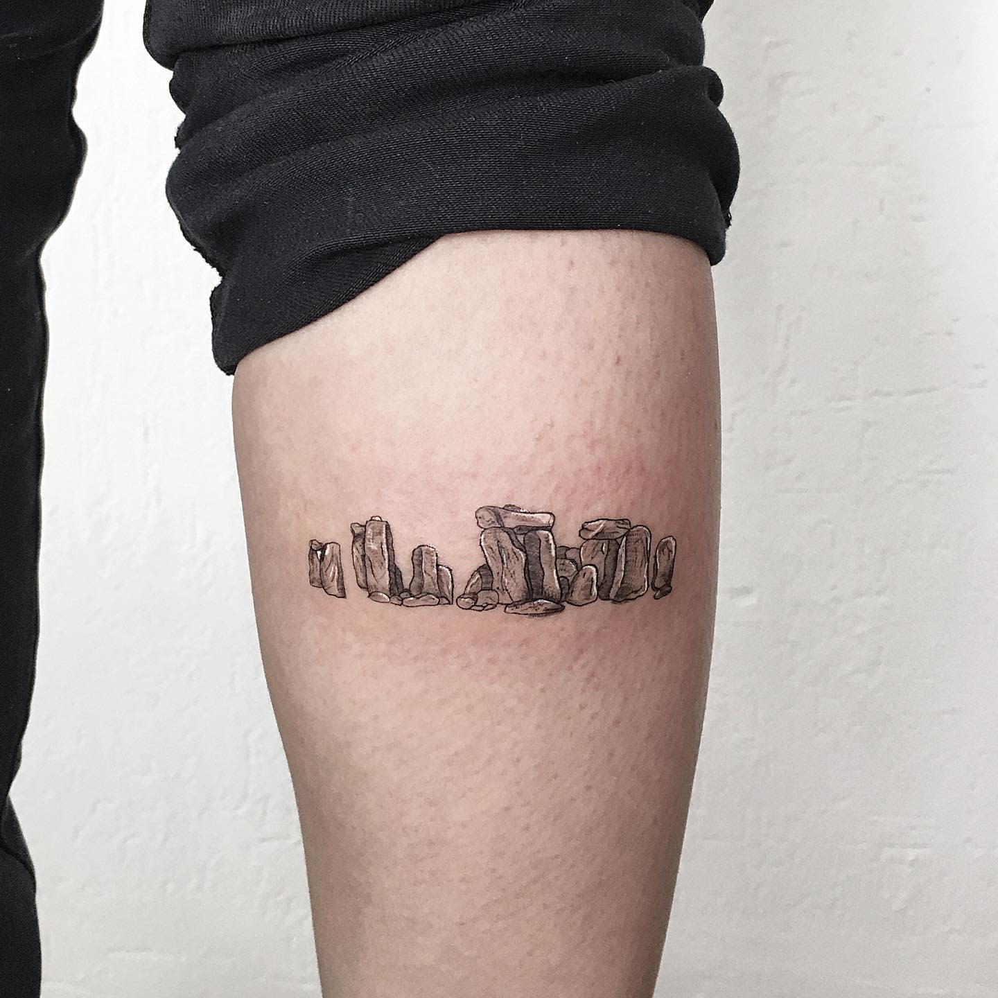 Small tattoo of Stonehenge on an inner arm, depicted in black ink with fine line details