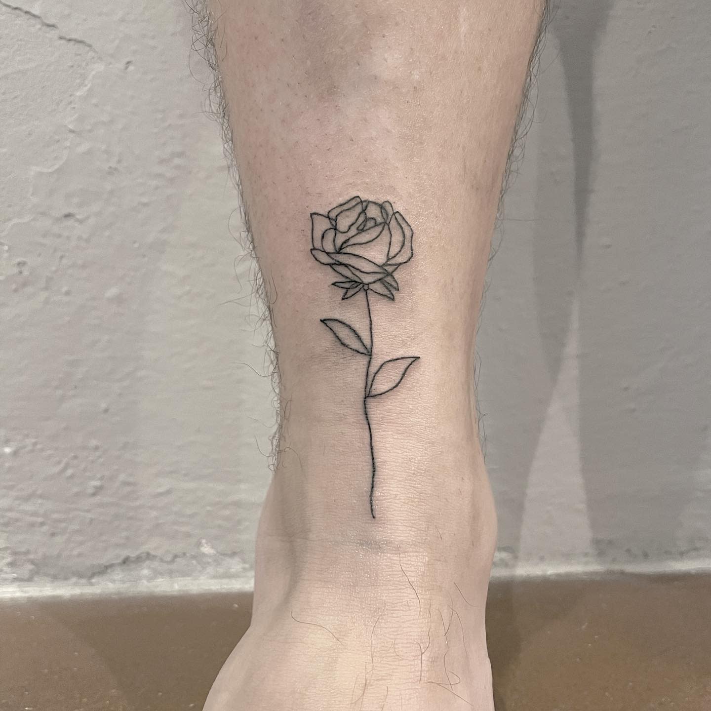 Fine line tattoo of a rose with detailed petals and leaves on someone's lower leg