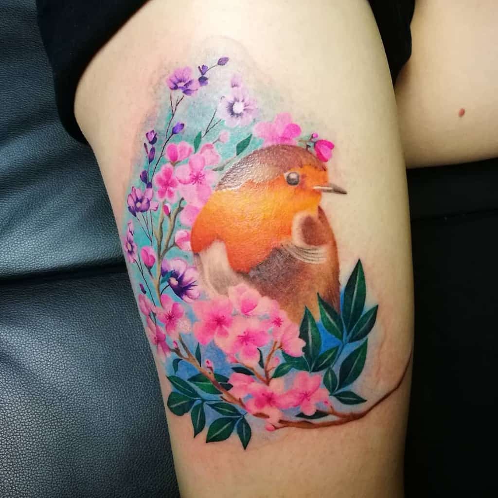 Vibrant watercolor-style tattoo of a robin surrounded by pink and violet blossoms and green leaves, with soft shading and a dreamy blue background effect