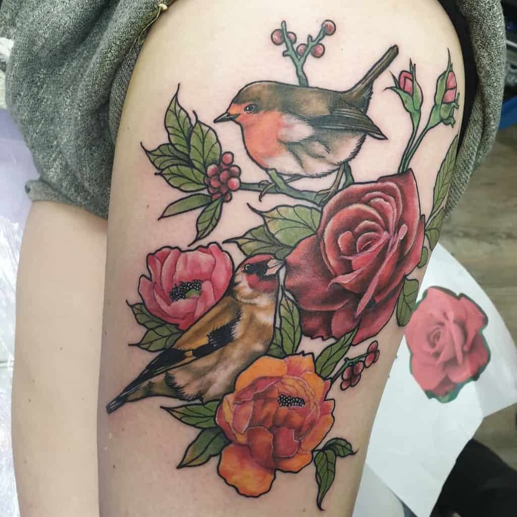 Colorful tattoo featuring a robin and a goldfinch perched among detailed roses, peonies, and leaves, with fine shading and vibrant tones