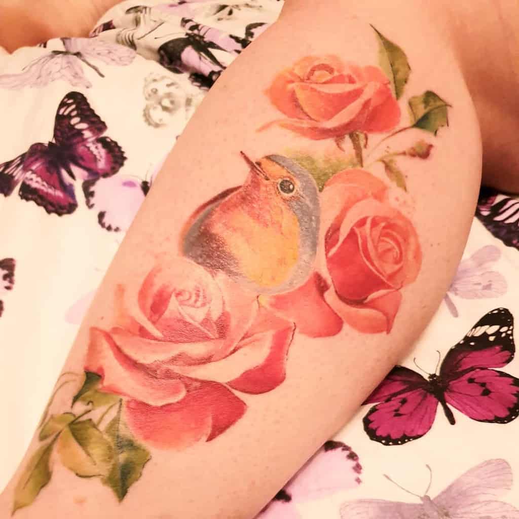 Soft watercolor-style tattoo of a robin surrounded by realistic red roses, featuring delicate shading and a natural blending effect