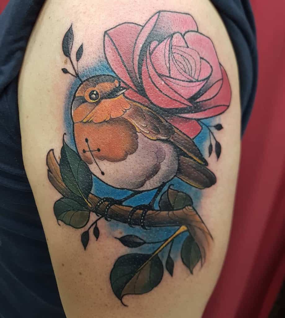 Colorful tattoo of a robin perched on a branch with a pink rose and leaves, against a blue backdrop