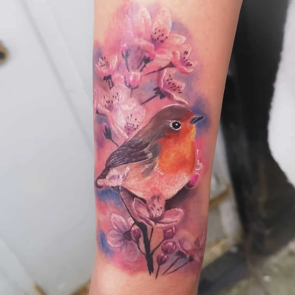 Colorful tattoo of a robin with an orange chest, perched on a branch, among pink cherry blossoms
