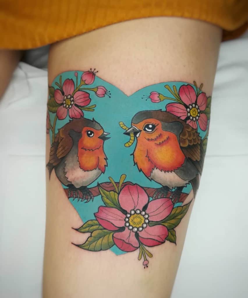Colorful tattoo of two robin perched on a branch with pink flowers and leaves, a heart-shaped blue background, one robin holds a worm in its beak