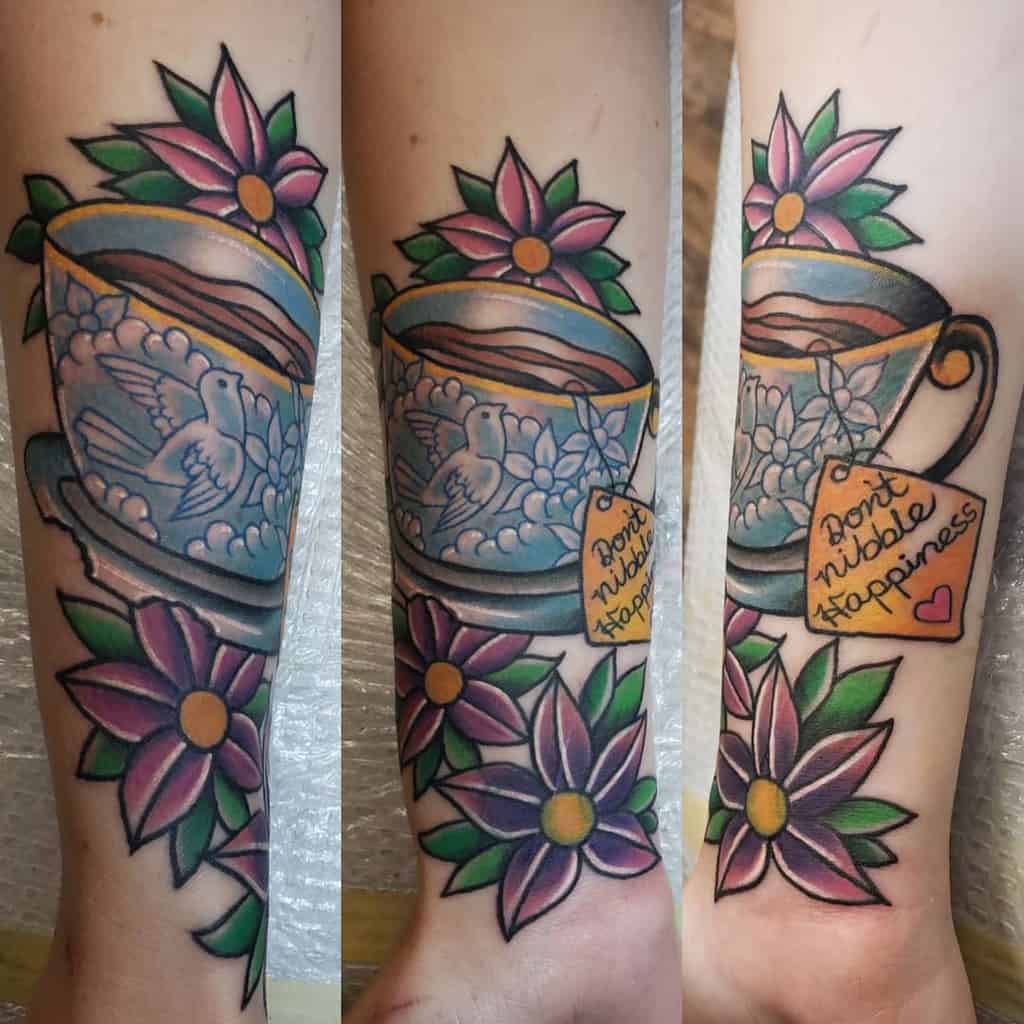 Colorful tattoo of a teacup with a dove, flowers, and a tag reading "Don't nibble happiness"