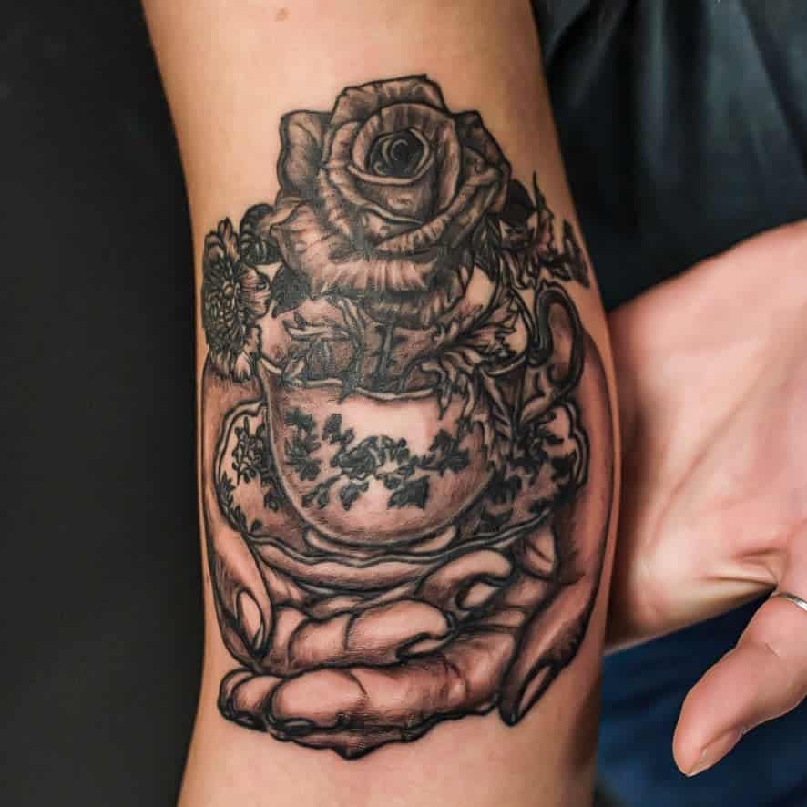 Tattoo of hands holding a floral teacup with a rose and other flowers blooming from it