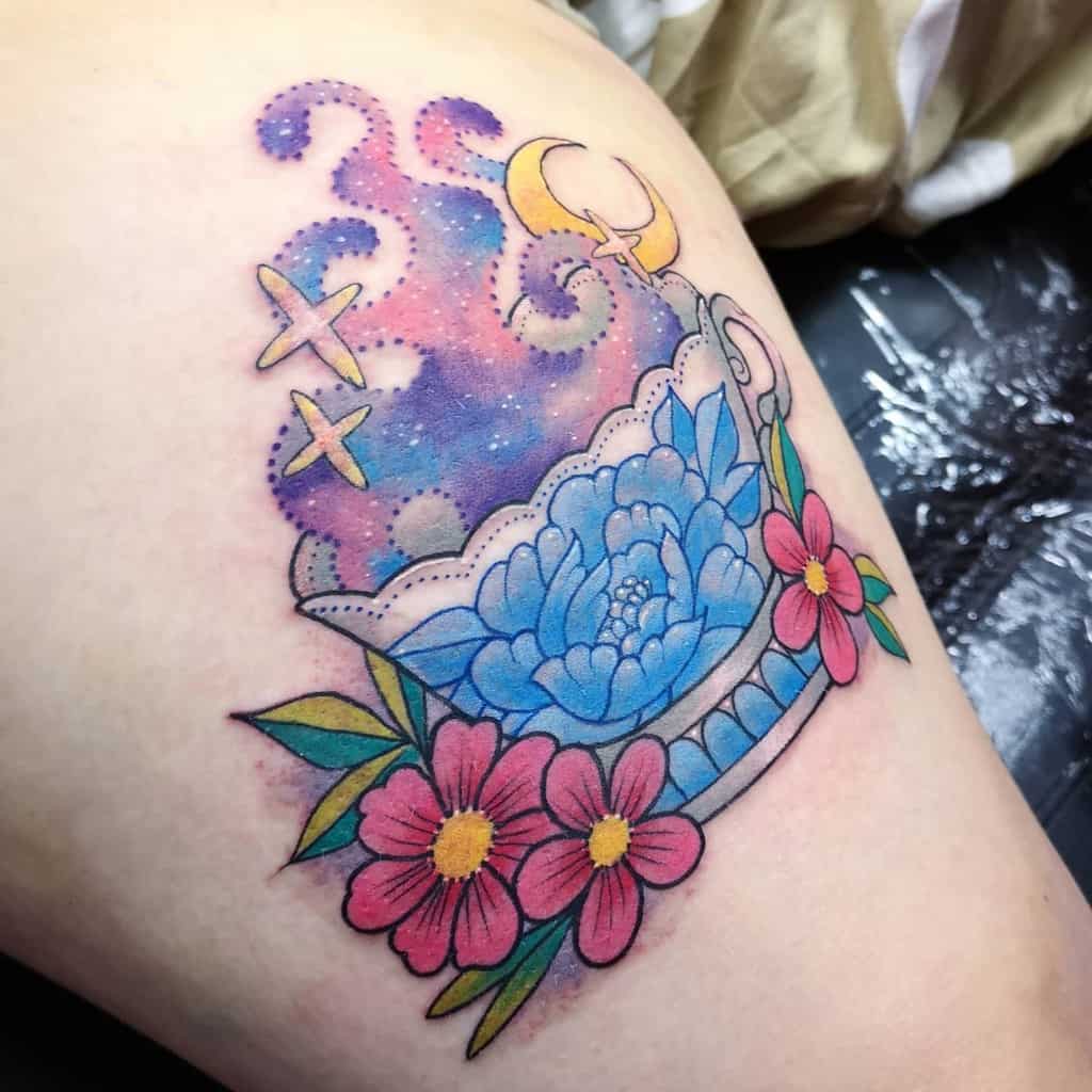 A colorful tattoo of a teacup with a blue flower, pink flowers, green leaves and swirling galaxy steam with stars and a crescent moon