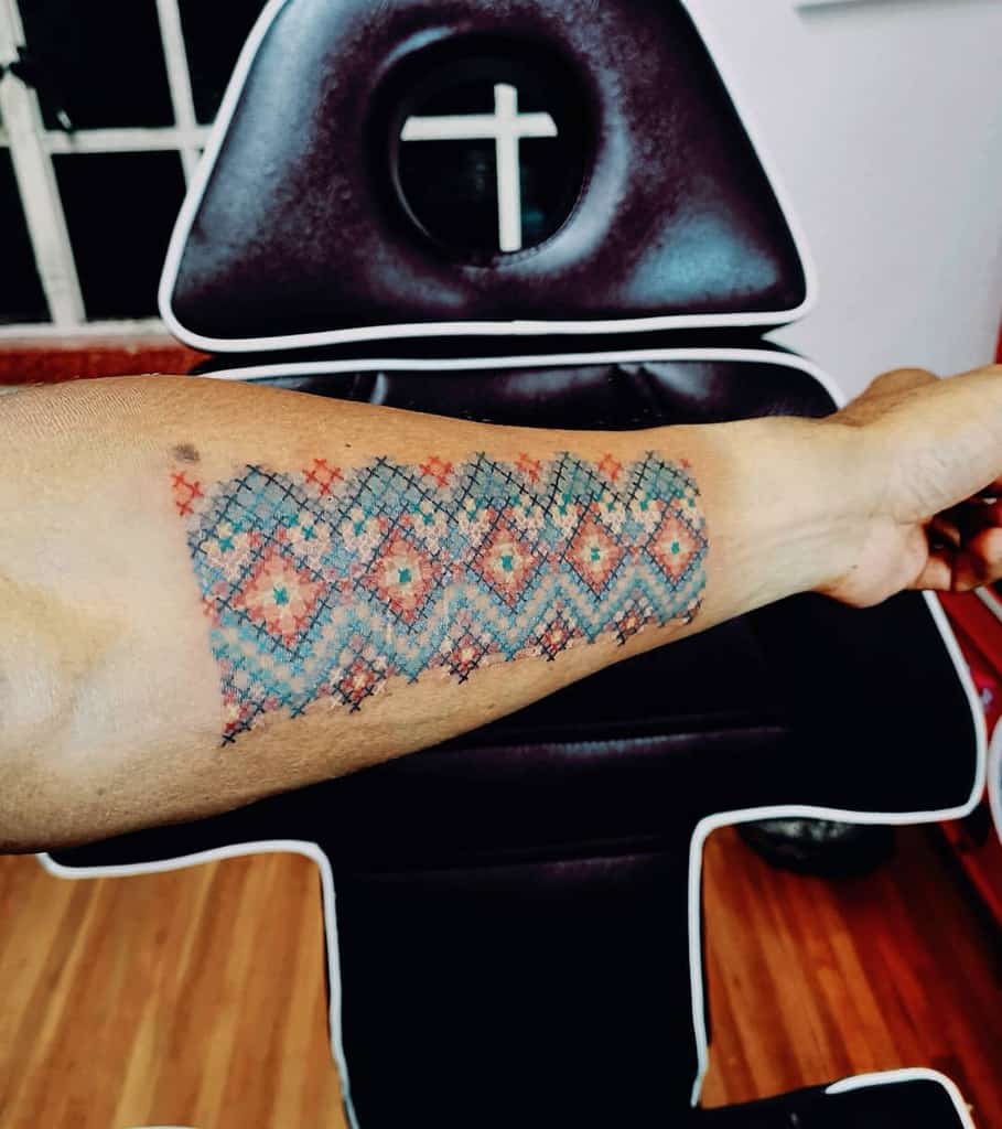 Intricate cross-stitch style tattoo of a geometric textile pattern in blue, red, and orange, mimicking traditional embroidery