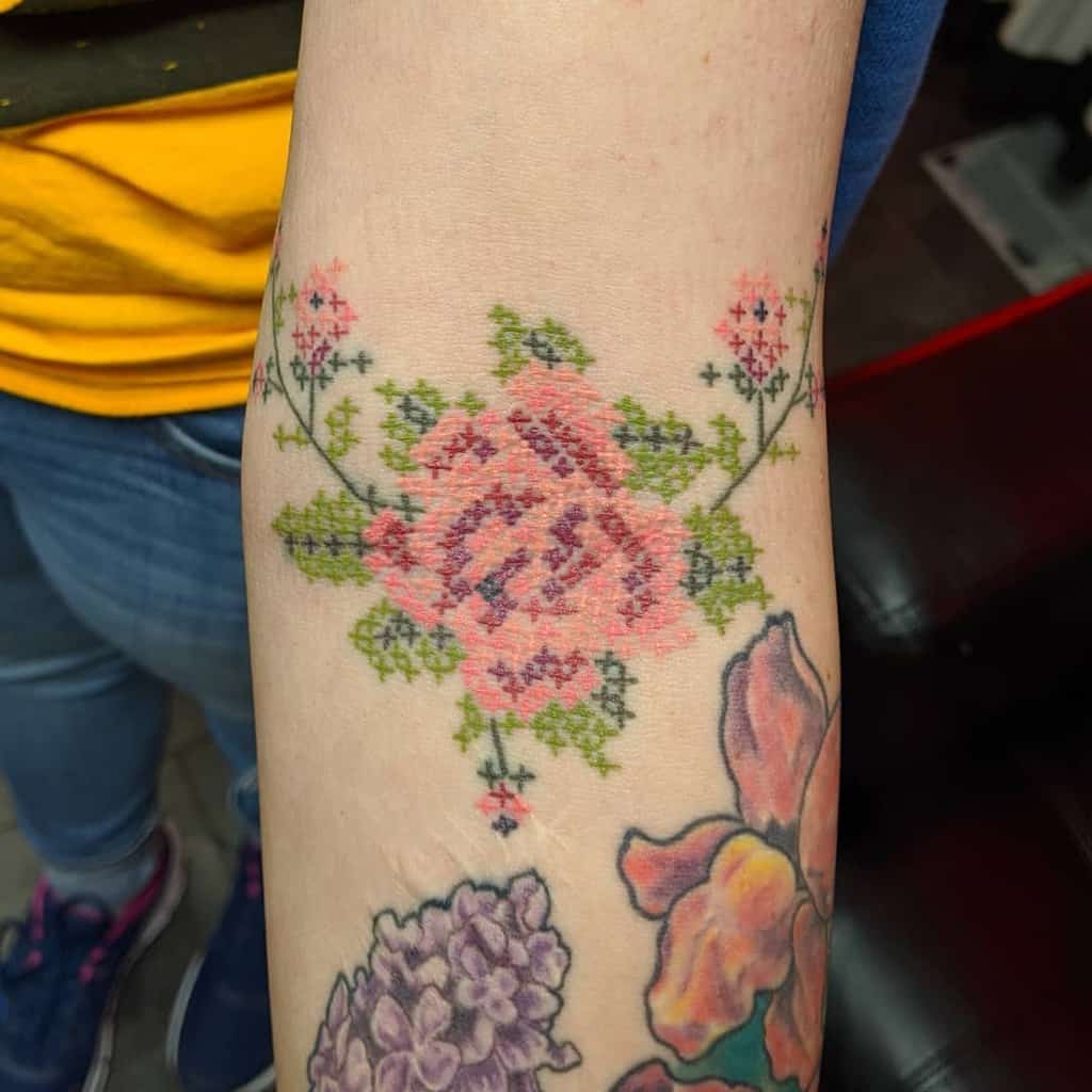 Beautiful cross-stitch style tattoo of a pink rose with green leaves and floral details, mimicking traditional embroidery