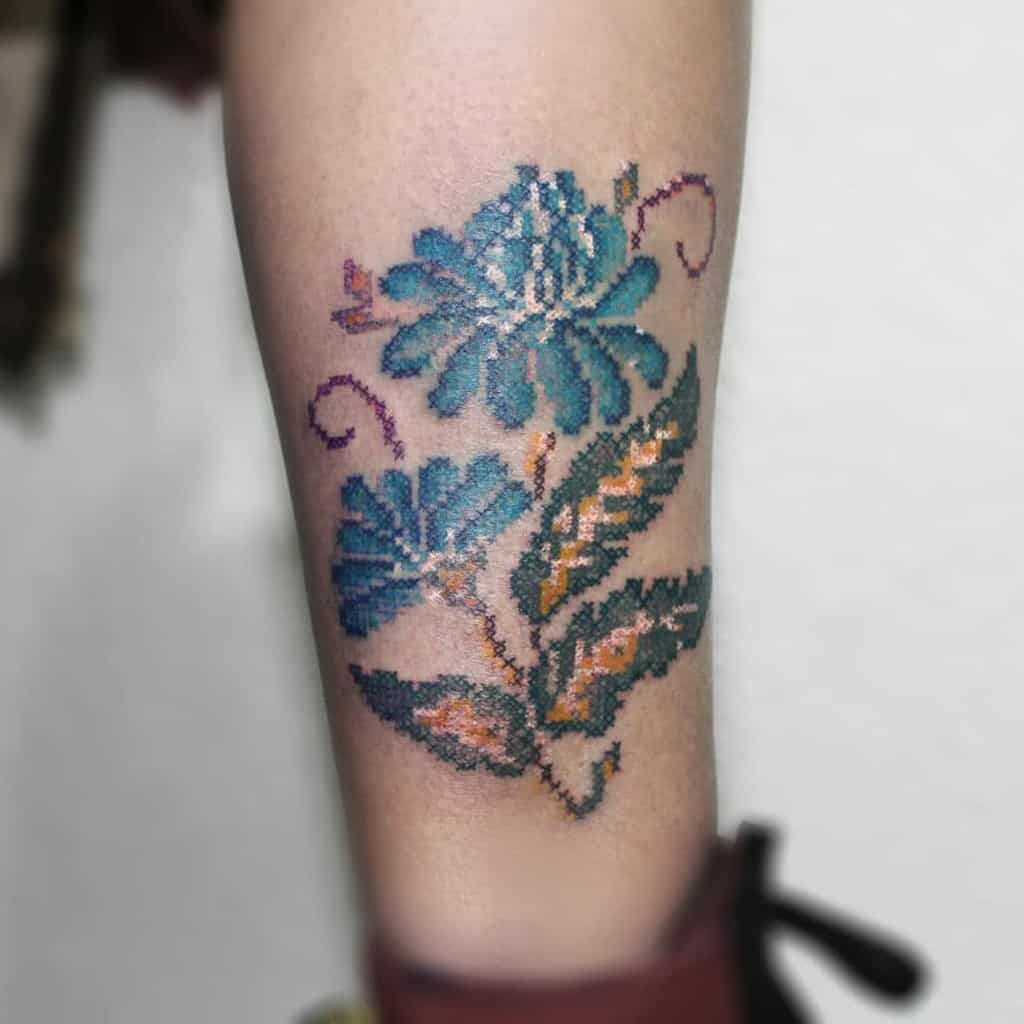 Cross-stitch tattoo of blue flowers with intricate leaves in warm tones, creating an embroidered effect for a unique and vintage-inspired look