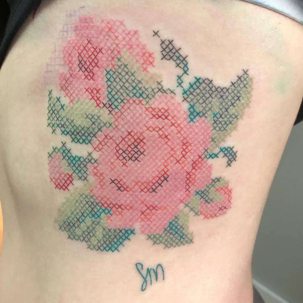 Cross-stitch tattoo of a pink rose with green leaves, designed to resemble embroidered floral patterns for a vintage aesthetic