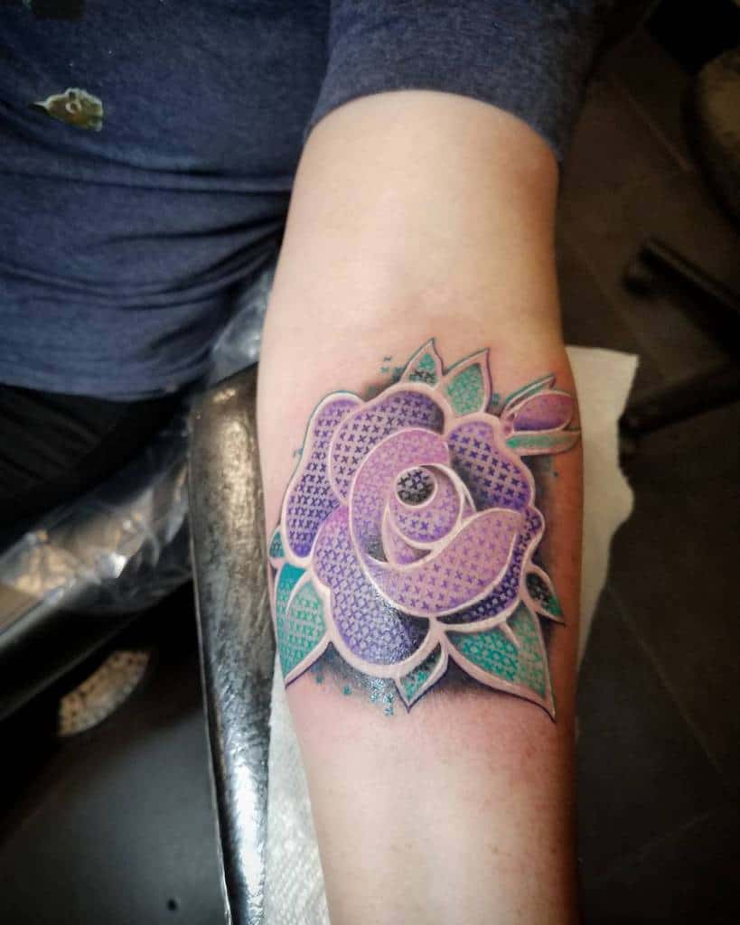 Cross-stitch tattoo of a vibrant purple and teal rose, designed with a 3D effect to resemble embroidered fabric
