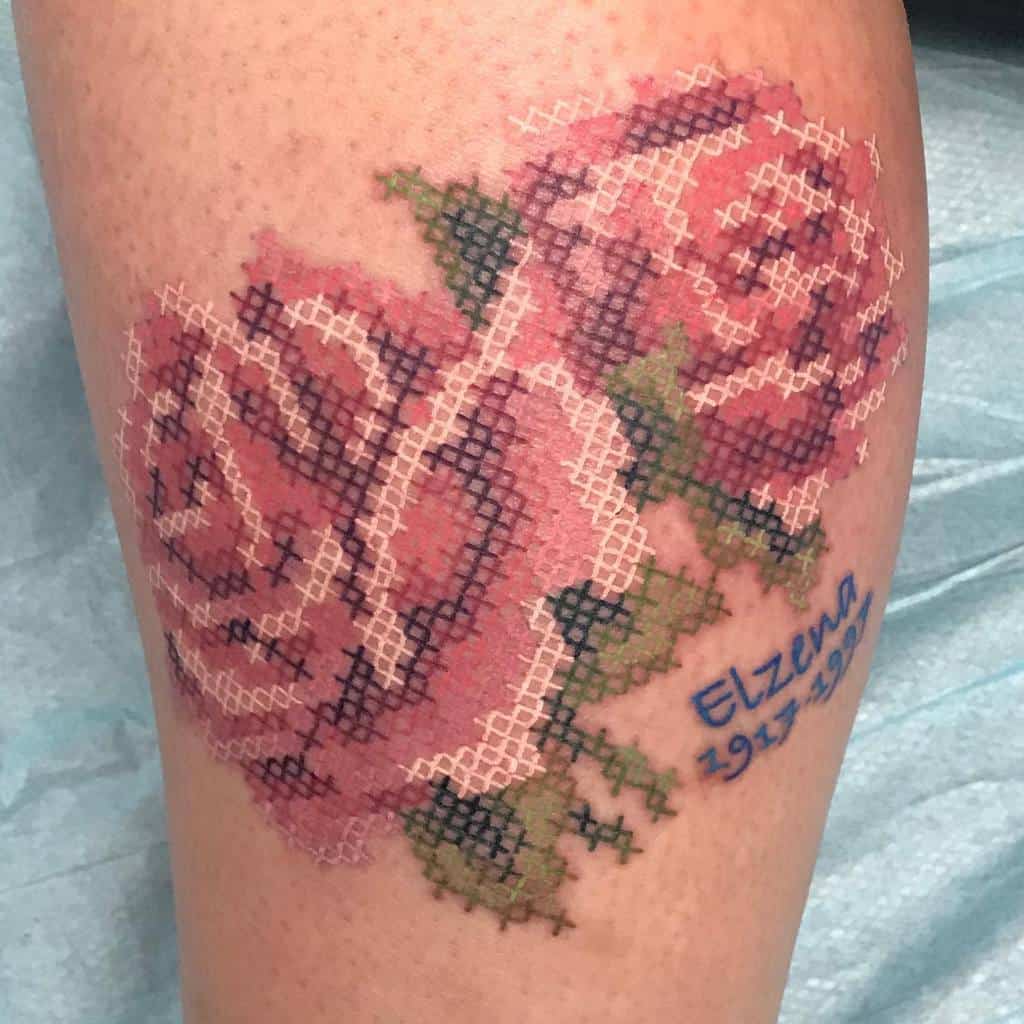 Cross-stitch tattoo of two pink roses with green leaves, designed to mimic embroidery, featuring a blue text 'Elzena 1917-1991'