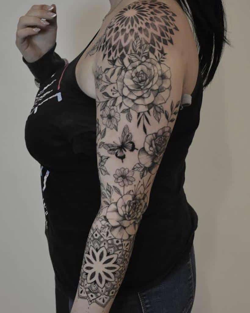 A black and grey floral sleeve tattoo featuring roses, butterflies, and geometric mandala patterns, covering the upper arm and forearm