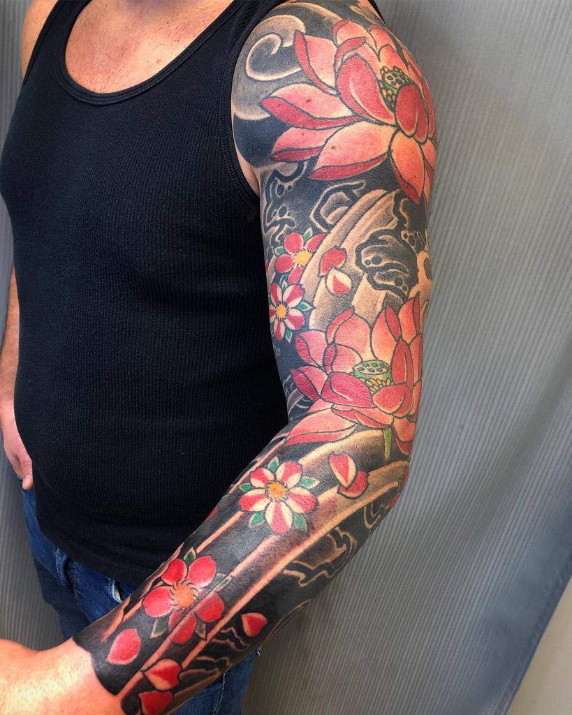 A vibrant full sleeve tattoo featuring red and pink lotus flowers with green accents against a dark background, wrapping around the arm