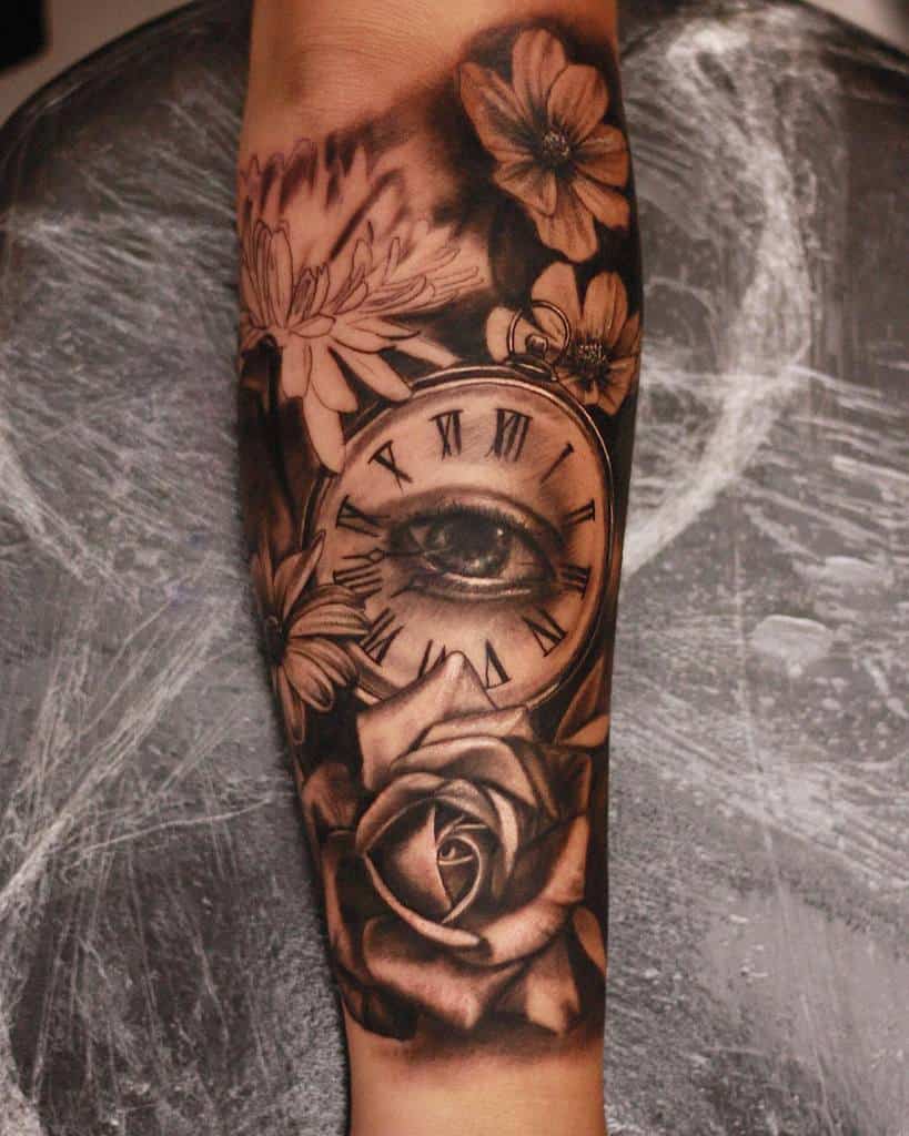 A black and grey forearm tattoo featuring an eye inside a clock with Roman numerals, surrounded by flowers and a detailed rose