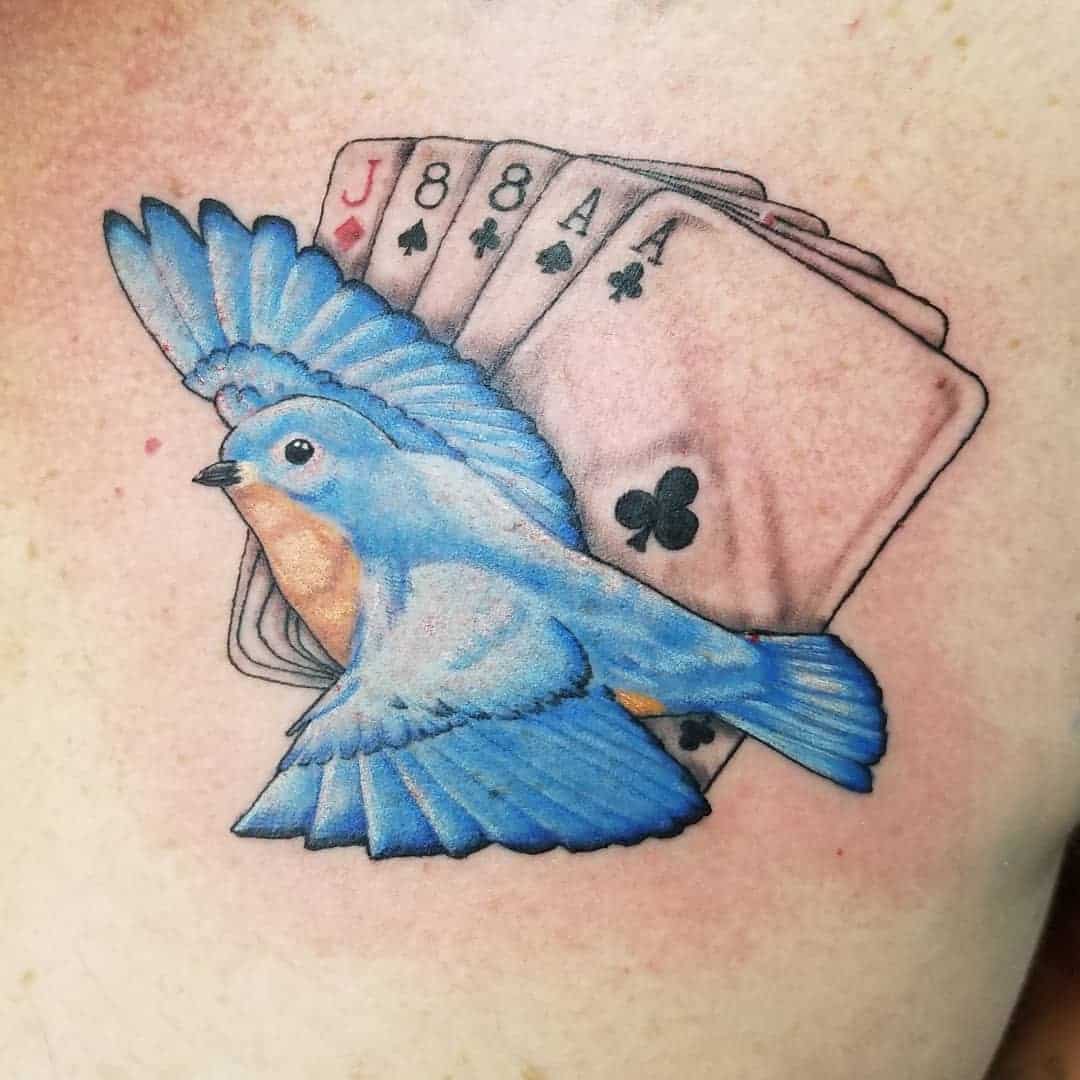 A tattoo of a bluebird flying in front of four playing cards: jack, eight of spades, eight, and ace of clubs