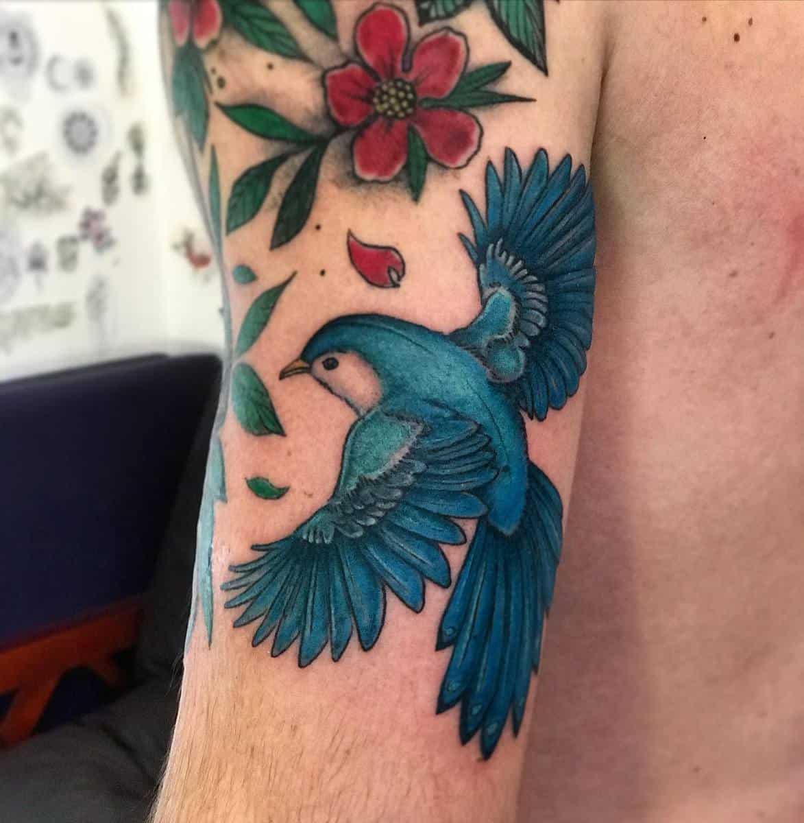Tattoo of a blue bird in flight surrounded by flowers and leaves on an upper arm