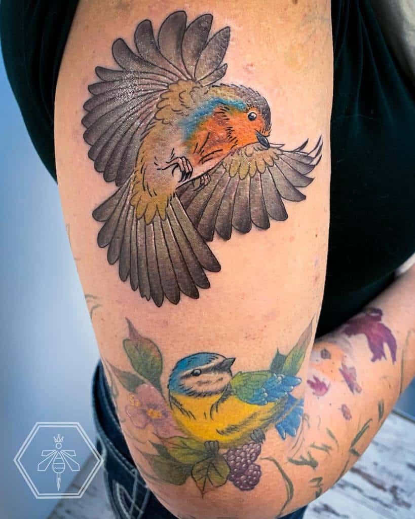 A colorful tattoo of two robin, a robin in flight above a robin perched on a branch with pink flower, leaves and berries