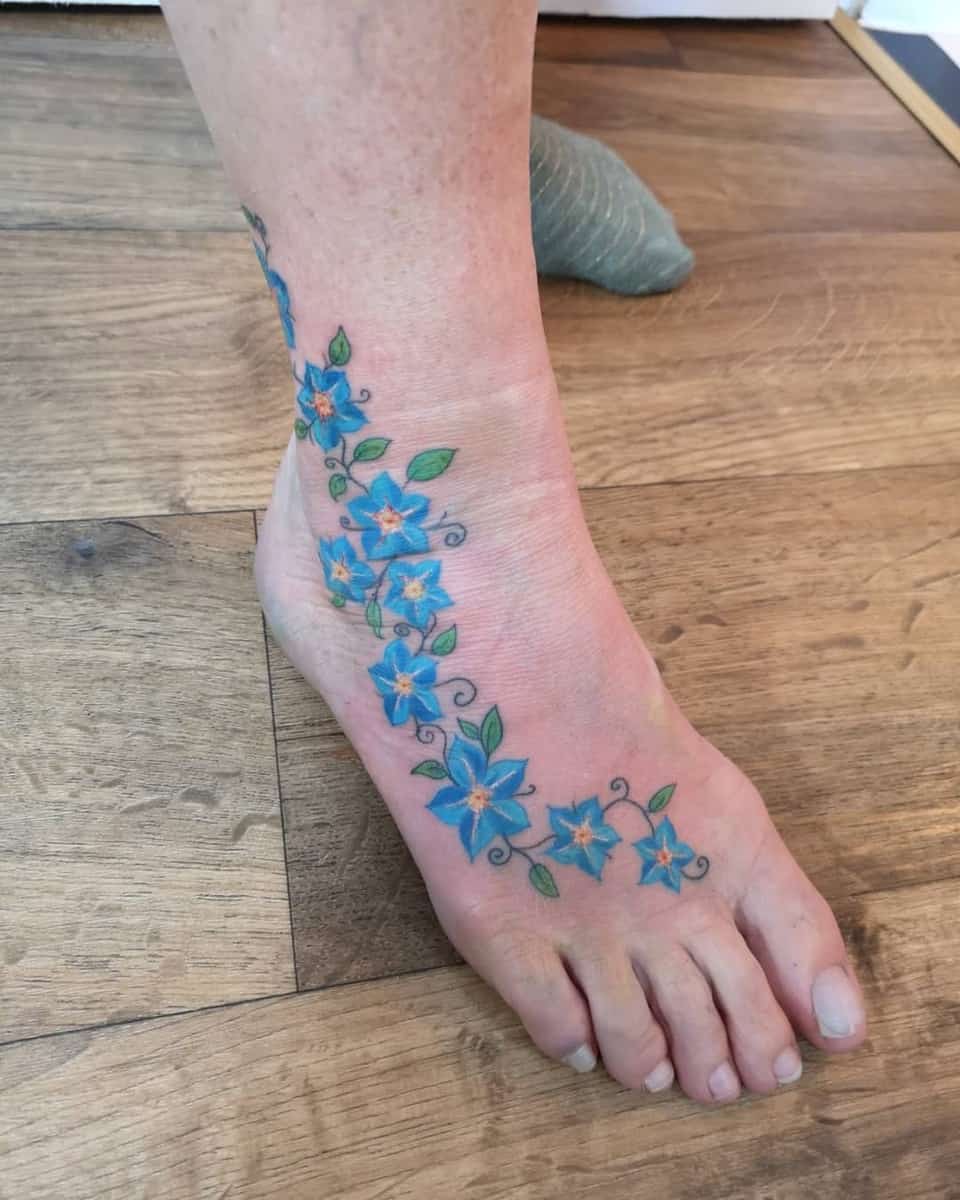 A foot tattoo featuring a vine of blue flowers with orange centers, extending from the ankle to the toes