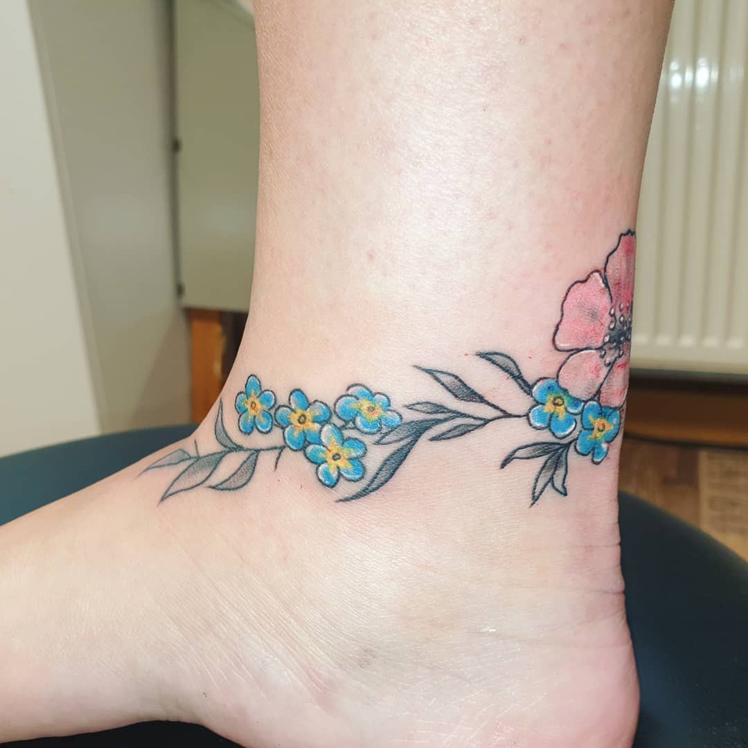 Ankle tattoo of blue and pink flowers with green leaves wraps around an ankle