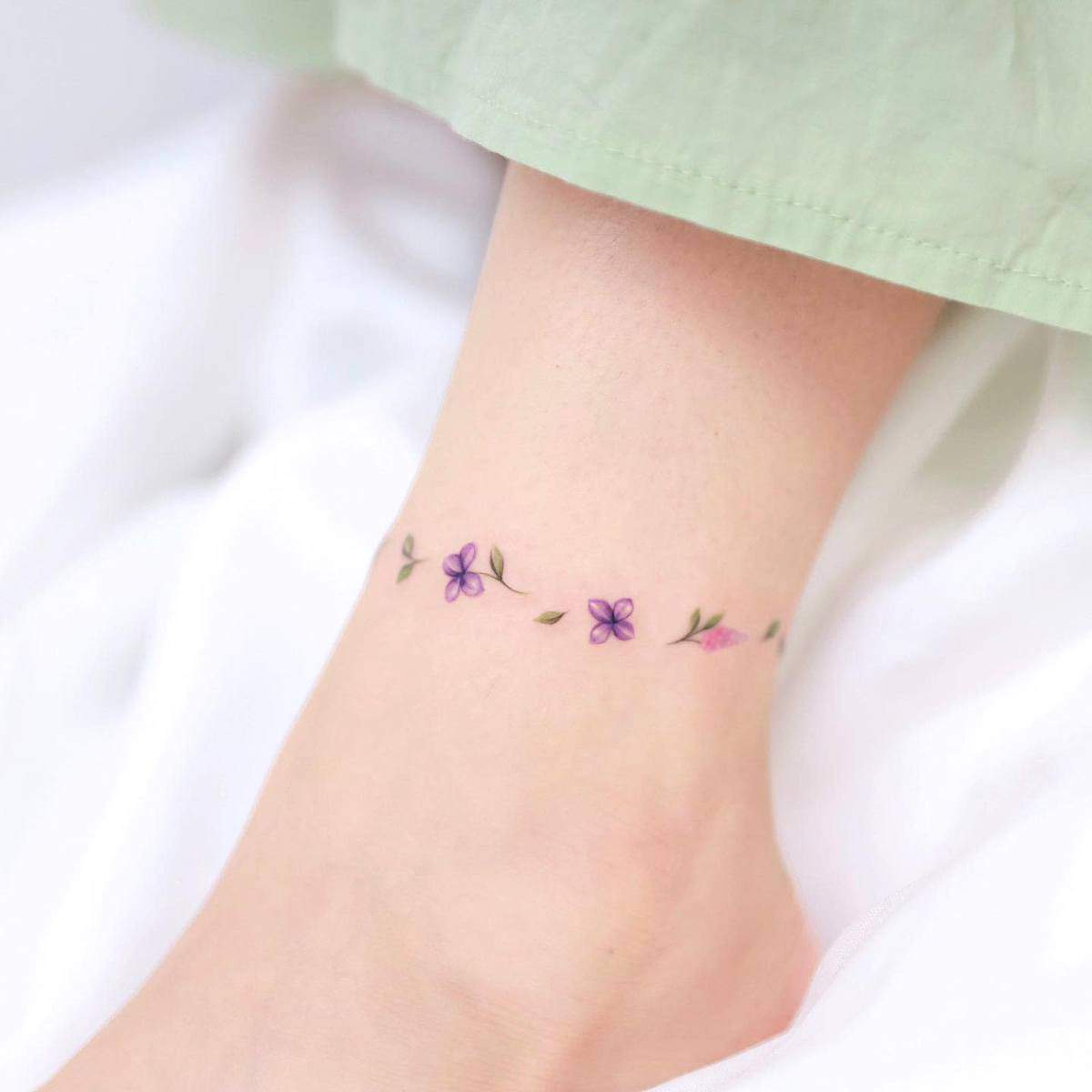 A delicate ankle tattoo with small purple and pink flowers and green leaves against light skin wearing pale green pants