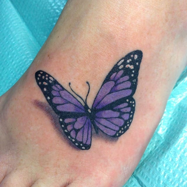 Purple butterfly tattoo on the top of a foot with a blue textured background